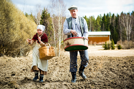 Mies ja tyttö kylvävät viljaa käsin vanhanajan vaatteissa, menossa on kylvönsiunaus