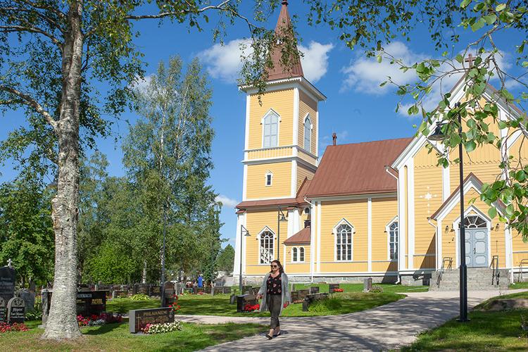 Nivalan keltainen kirkko taustalla kesäisessä auringonpaisteessa ja etualalla kävelee nainen.